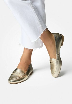 Metallic gold Loafer mit glänzender Oberfläche, die über eine genähte Detailverarbeitung an der Vorderseite und einen niedrigen, abgerundeten Absatz verfügen. Getragen mit weißen, ausgefransten Jeans.
