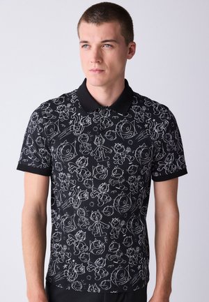 Tânăr purtând un tricou polo negru cu imprimeu alb cu pisici desenate, stând în picioare în fața unui fundal simplu, deschis la culoare.