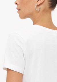 Witte T-shirt met korte mouwen en opgerolde mouwen, met een zachte textuur. Een zilveren ringoorbel is zichtbaar aan het oor van het model.