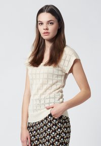 Virkat tröja i ljus crème med ett rutmönster; korta ärmar och en texturerad yta. Kombinerad med mönstrade svarta och gula byxor.