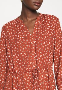 Vestido color óxido con un estampado floral, mangas largas, escote en pico y cintura con cordón; confeccionado en una tela ligera y fluida.