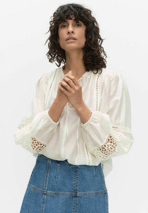 Vrouw met krullend haar, gekleed in een witte blouse met kanten details en pofmouwen, gecombineerd met een blauwe spijkerrok, handen in elkaar gevouwen.