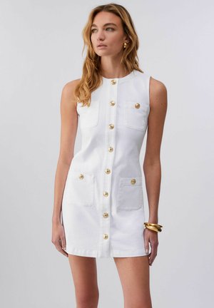 Donna che indossa un vestito bianco senza maniche con bottoni dorati e tasche, accessoriato con orecchini e braccialetti dorati.