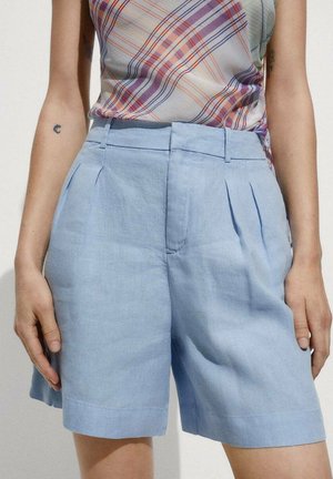 Light blue linen shorts with pleats, a smooth texture, and a waistband. Styled with a plaid top for contrast. Tailored mid-thigh length.