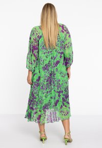 Blumenmusterkleid in Grün und Lila, mit transparenter Stoff, langen Ärmeln und einer fließenden Silhouette, die bis zur Mitte der Waden reicht.