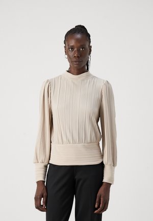 Blouse plissée beige avec un col montant, des manches longues froncées et un ourlet texturé, associée à un pantalon noir ajusté.