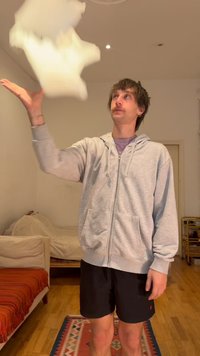 Jeune homme en sweat à capuche gris et short noir lançant un oreiller blanc dans un salon avec un sol en bois et un tapis coloré.