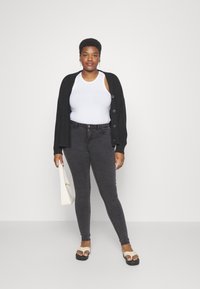 Cárdigan negro de punto, top blanco sin mangas, jeans ajustados gris oscuro, sandalias planas color tostado y bolso blanco. Diseño simple y ajustado con botones.