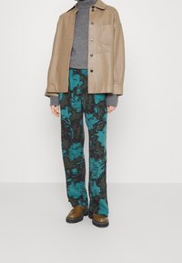 Tan leren jas met een borstzak, grijze gebreide trui en teal broek met bloemenpatroon. Bruine stevige schoenen maken de look compleet.