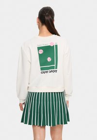 Sweatshirt crème avec un graphisme représentant un court de tennis vert et des balles de tennis roses. Associé à une jupe plissée rayée verte et blanche.
