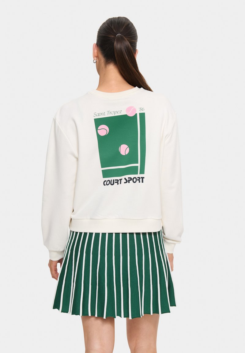 Sweatshirt crème avec un graphisme représentant un court de tennis vert et des balles de tennis roses. Associé à une jupe plissée rayée verte et blanche.