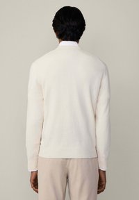 Mann mit kurzen schwarzen Haaren, der einen weißen Pullover über einem weißen Hemd und beigen Hosen trägt, mit dem Rücken zu einem neutralen Hintergrund. 