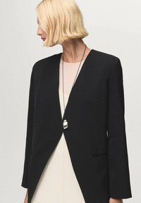 Blazer nero strutturato con scollatura a V e maniche lunghe. Caratterizzato da un design minimalista e da una texture liscia, indossato sopra una maglietta di colore chiaro.