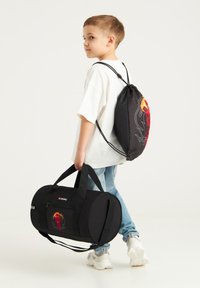 Czarny plecak na sznurki z czerwoną i żółtą grafiką oraz czarna torba sportowa z przednią kieszenią na suwak, trzymane przez dziecko. Biała koszulka, niebieskie dżinsy.