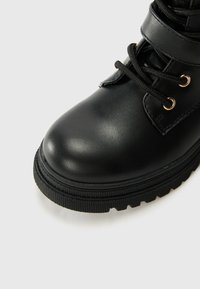 Svarta läderankelboots med rund tå, texturerad gummisula och ögla i guld för snörningen. Matt yta utan synliga mönster.