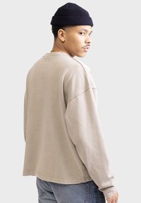 Beige Langarmshirt mit überschnittenen Schultern, rundem Ausschnitt und lockerer Passform. Kombiniert mit blauen Jeans und einer dunklen Strickmütze.