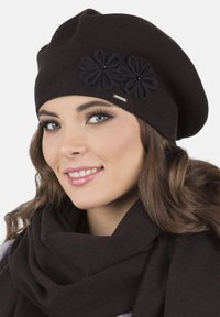 Černý pletený baret s třemi látkovými květinami a korálkovými akcenty, s kovovou etiketou, nošený s černým šátkem. Detail obličeje.