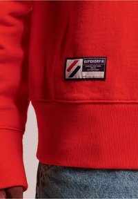 Červená mikina s vroubkovanými manžetami a lemem, s obdélníkovou nášivkou s nápisem "SUPERDRY" a geometrickými vzory.