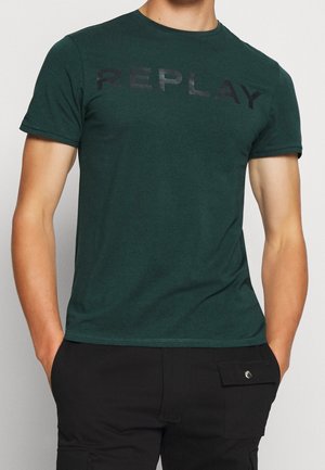 Homme portant un T-shirt à manches courtes vert foncé avec "REPLAY" imprimé sur la poitrine et un pantalon noir avec une poche à bouton.