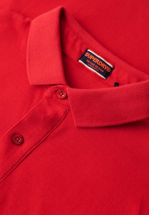 Czerwona koszulka polo z teksturowanej tkaniny, z klasycznym kołnierzykiem, dwoma guzikami i metką Superdry przy szyi.
