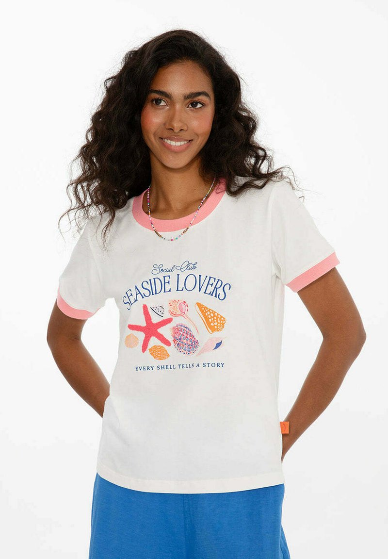 Žena s kudrnatými vlasy, oblečená v bílo-růžovém tričku s potiskem mušlí a hvězdic a modrých kalhotách, usmívající se s rukama za zády.