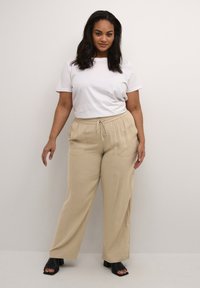 Pantalons beiges à jambes larges avec une taille élastique et un cordon de serrage. Associés à un t-shirt blanc uni et des sandales noires à talons carrés.