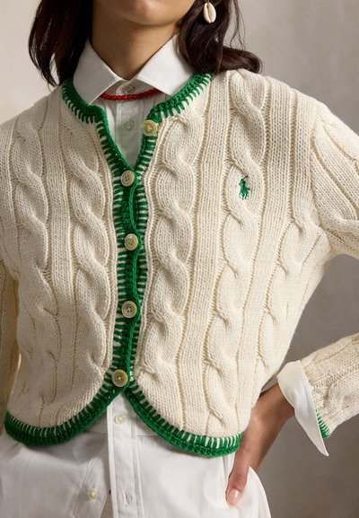 Sieviete valkā krēmkrāsas adītu kardiganu ar zaļu apmali un pogām virs baltas krekla ar apkakli, aksesuāri papildināti ar sarkanām krellēm un gliemežvāku auskariem.