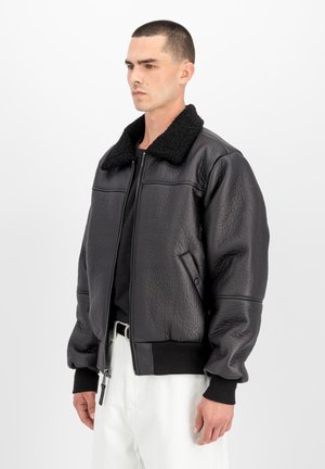 Man wearing a black textured leather jacket with a black shearling collar, black shirt, and white pants against a plain white background.