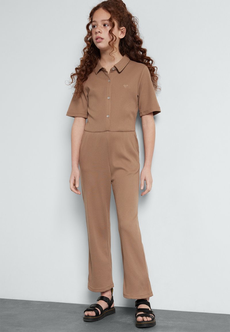 Jumpsuit marron à côtes avec des manches courtes, fermeture à boutons et pantalons évasés. Assorti à des sandales noires avec de larges lanières et une semelle épaisse.