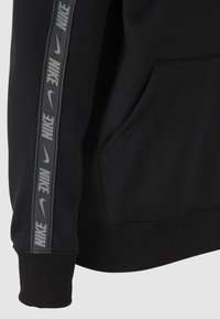 Sudadera negra con una textura suave, cinta con el logotipo prominente de Nike en la manga izquierda y un bolsillo tipo canguro en la parte frontal.