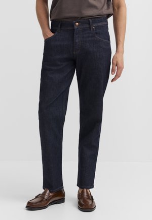 Mann trägt dunkelblaue Jeans mit geradem Bein, braune Leder-Loafer und ein eingestecktes graues Hemd, steht mit einer Hand in der Tasche.