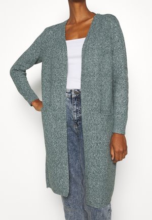 Długi szary dziergany kardigan z otwartym przodem i dwoma kieszeniami z przodu, noszony na białym topie i niebieskich dżinsach z wysokim stanem.
