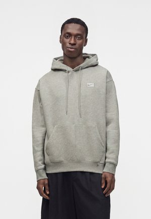 Άνδρας που φορά ανοιχτό γκρι φούτερ Nike με μπροστινή τσέπη και μαύρο παντελόνι, στέκεται μπροστά σε απλό λευκό φόντο.