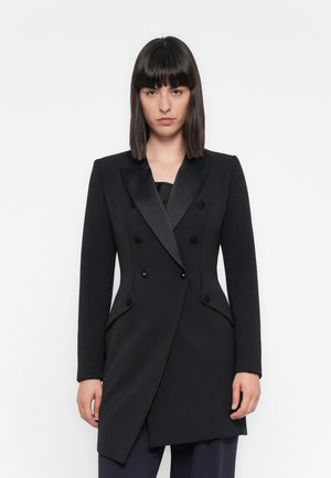 Woman with straight dark hair wearing a fitted black double-breasted blazer with satin lapels and asymmetrical hem against a white background.