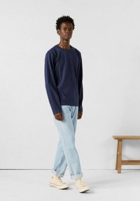 Marineblauwe lange mouwen shirt, katoenen stof, ontspannen pasvorm. Gecombineerd met lichtblauwe opgerolde jeans en beige hoge sneakers. Effen achtergrond.