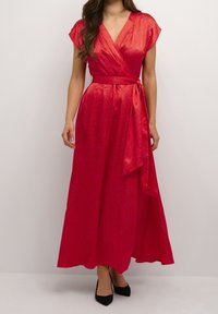 Kvinna i en livlig röd omslagklänning med subtilt mönster, korta ärmar, knytning i midjan och svarta spetsiga klackar, stående framför en neutral bakgrund.