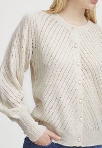 Cardigan beige clair avec des rayures verticales et une texture en tricot ajouré. Présente un col rond et des boutons en perles sur le devant. Manches à bords côtes.