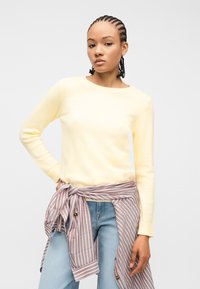 Jeune femme aux cheveux tressés portant un pull jaune pâle et un jean bleu clair, avec une chemise rayée nouée autour de la taille.
