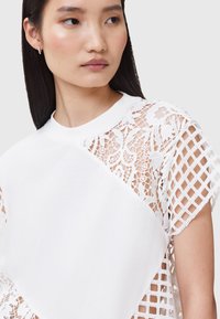White t-shirt featuring detailed lace and open grid pattern on sleeves, with a round neckline and soft texture, creating a layered effect.