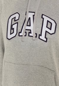 Sudadera gris con capucha, que presenta grandes letras bordadas "GAP" en blanco y marino. Incluye un bolsillo frontal tipo pouch y cordones ajustables.