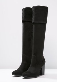Bottes hauteur genou en suède noir avec revers pliés, fermetures éclair latérales, bouts pointus et talons hauts épais, posées sur une surface blanche.
