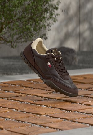 Zapatilla deportiva marrón oscuro con interior beige y suela negra flotando sobre baldosas de madera al aire libre cerca de una pared de hormigón y follaje verde.
