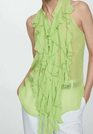 Lichtgroene doorschijnende blouse met verticale ruches aan de voorkant, knoopsluiting en een losse pasvorm. Gecombineerd met witte broek.