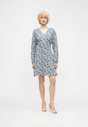 Femme aux cheveux blonds courts et bouclés portant une robe à manches longues bleu et blanc à fleurs et des sandales à talons beiges, debout devant un fond uni.