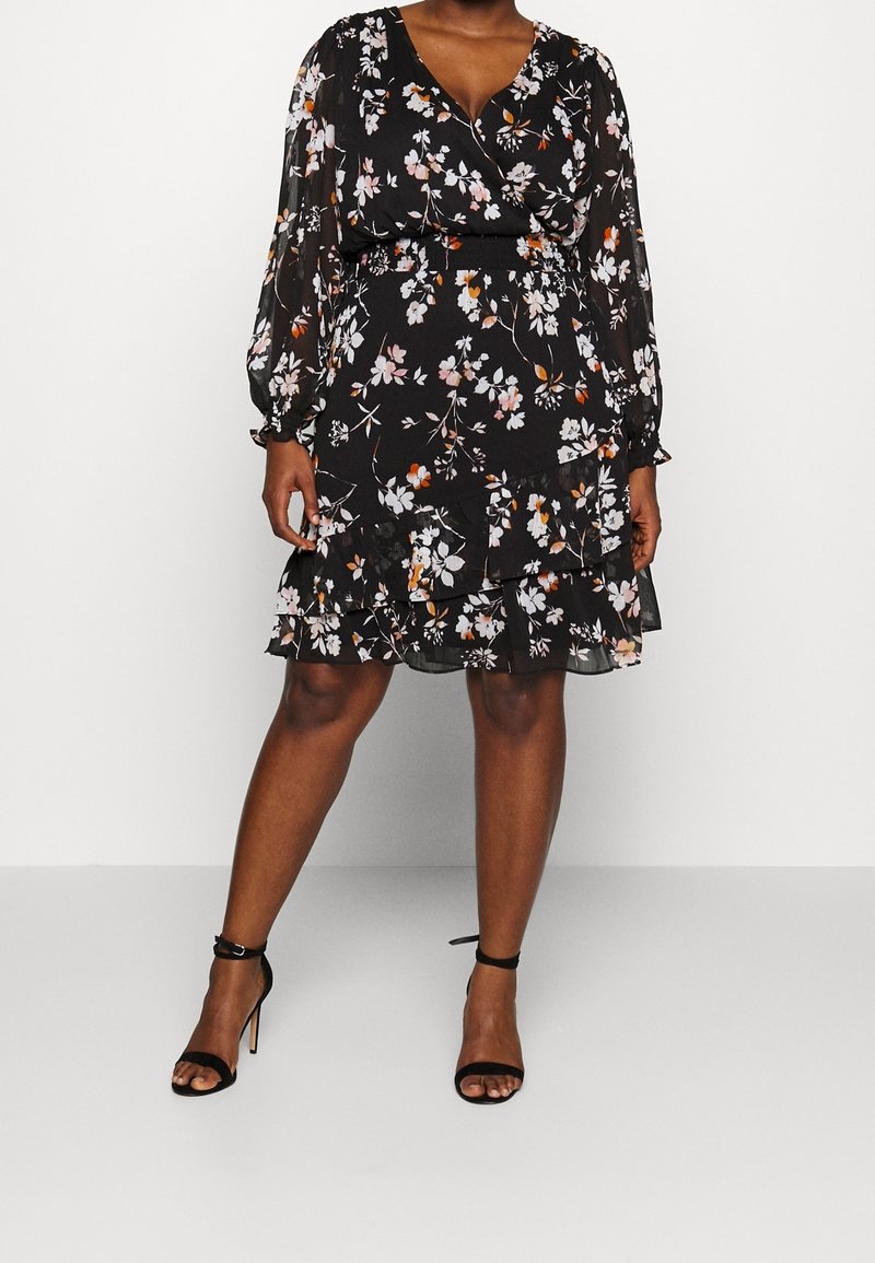 Robe noire à fleurs avec manches longues transparentes, décolleté en V et jupe à volants à étages. Présente des motifs floraux roses, blancs et orange. Portée avec des talons noirs.