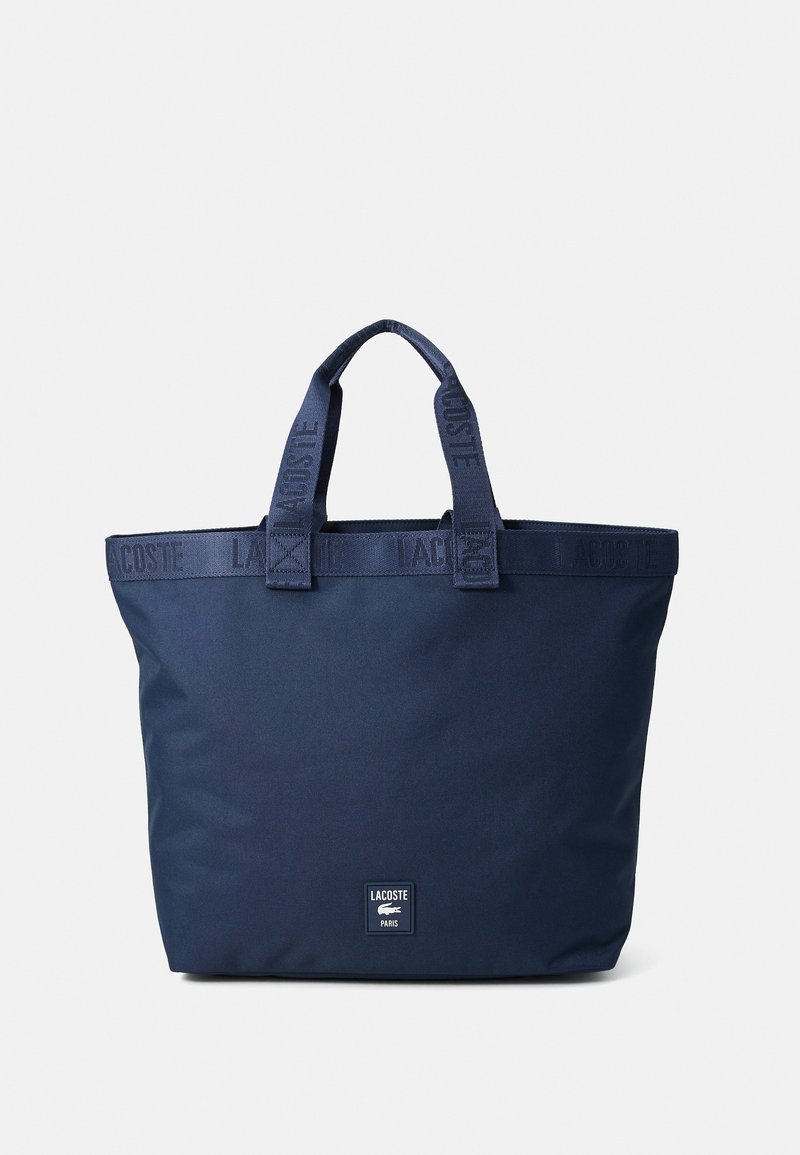 Mørkeblå tote bag laget av slitesterkt stoff, med to håndtak på toppen og et logo-patch på forsiden. Teksturert stroppdetalj forbedrer designet.