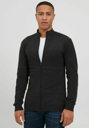 Black zip-up jacket with high collar, long sleeves, ribbed cuffs, and hem. Smooth texture with a fitted design, worn over a white shirt.