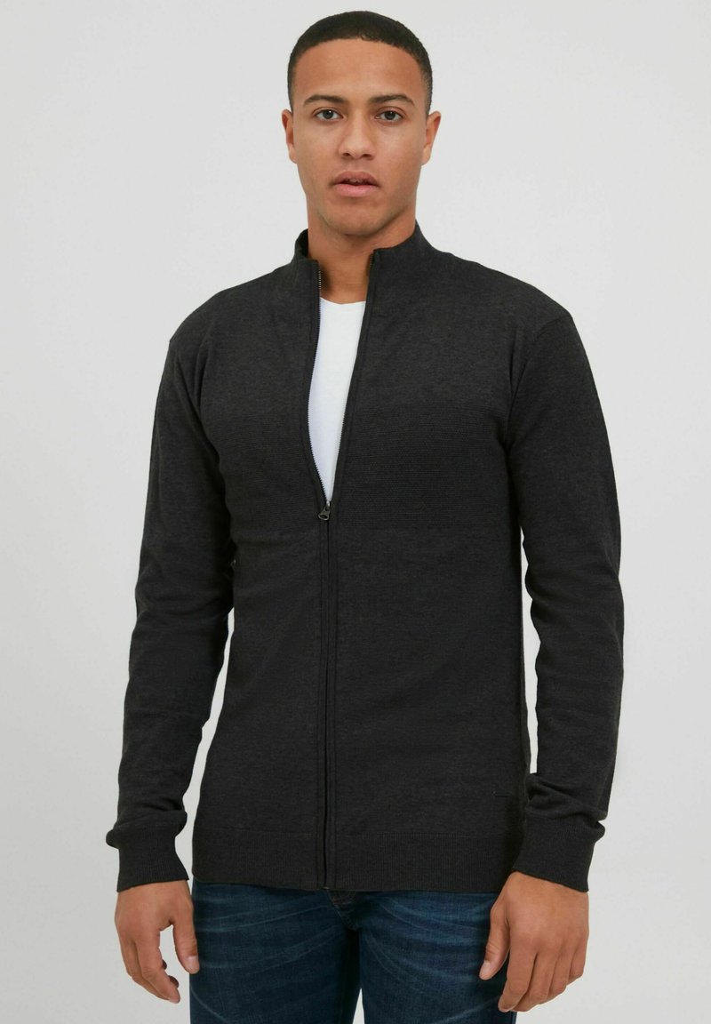 Svart zip-up jacka med hög krage, långa ärmar, ribbade mudd och nederkant. Slät textur med en åtsittande design, bärs över en vit skjorta.