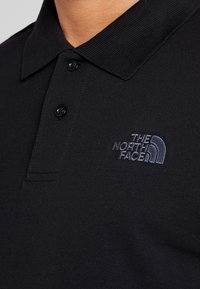 Černé polo tričko vyrobené z strukturované tkaniny s tříknoflíkovou légou. Na levé straně je tmavě vyšívané logo.
