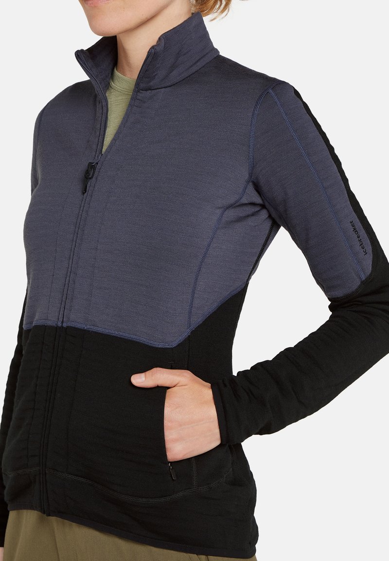 Frau trägt eine zweifarbige Zip-Jacke mit einer Hand in der Seitentasche, mit der oberen Hälfte in Dunkelblau und der unteren Hälfte in Schwarz.